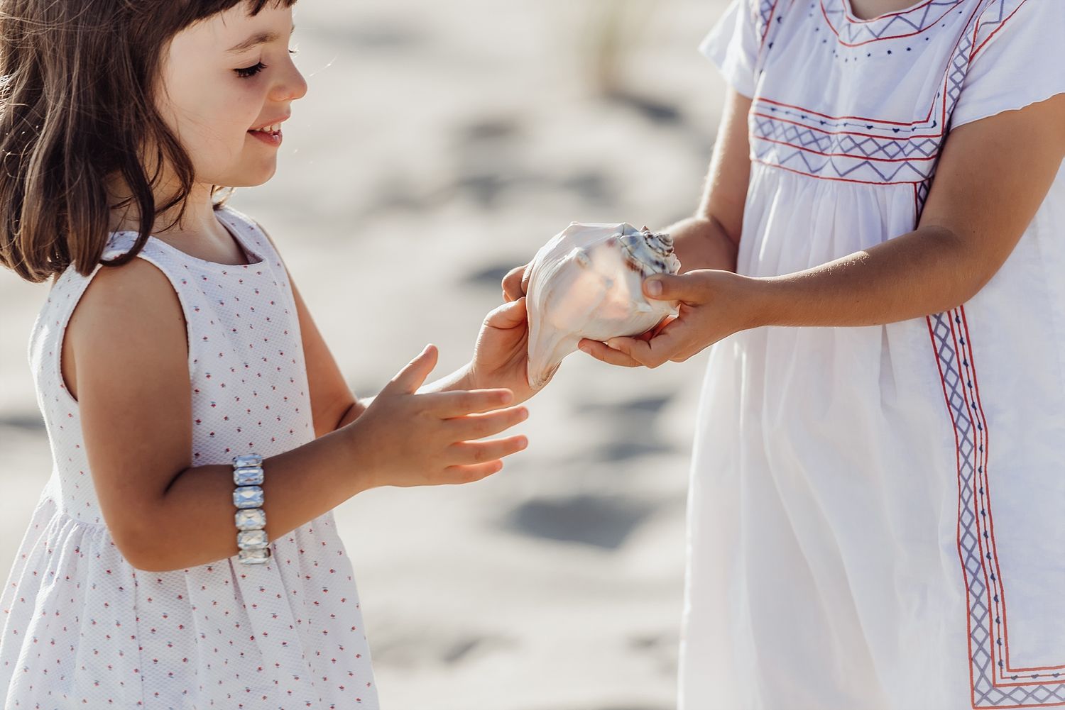 Two people in white dresses examine a small object together on a sunny beach.