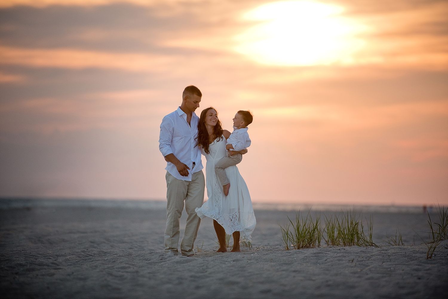 Family of three stands together on beach during golden sunset hour.