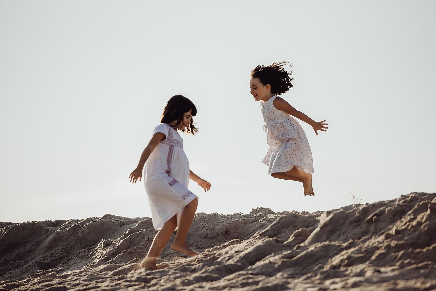 Two people in white dresses leap joyfully on sand dunes against bright sky.