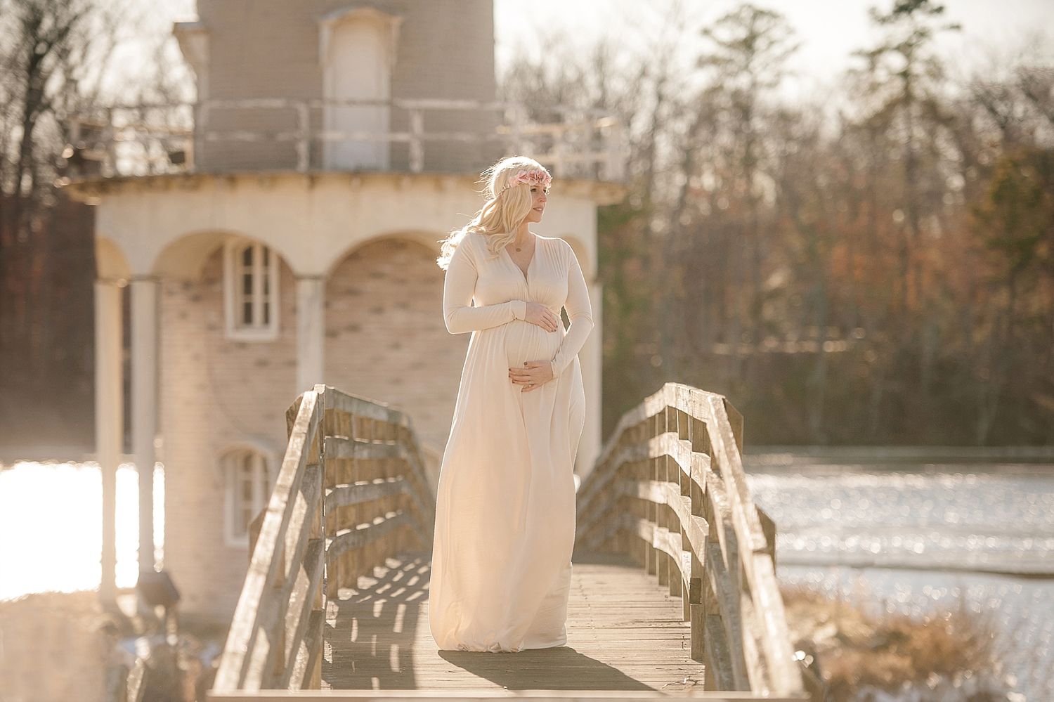 A person in flowing white dress stands on wooden bridge near classical building at sunset.