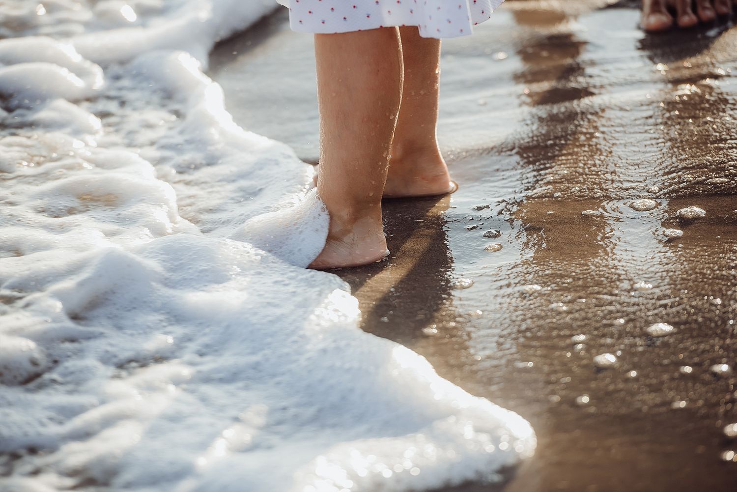 Feet standing at the edge of ocean waves and foam on a sandy beach.