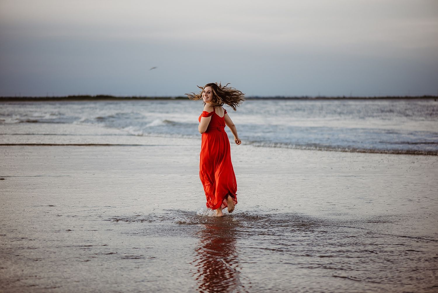 Woman in flowing red dress walks along beach shore at dusk.