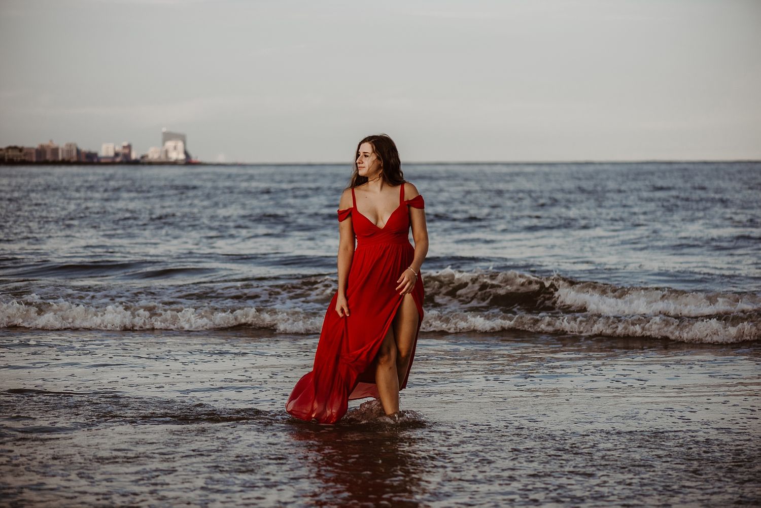 A red dress flows in the ocean breeze during a beachside photoshoot at dusk.