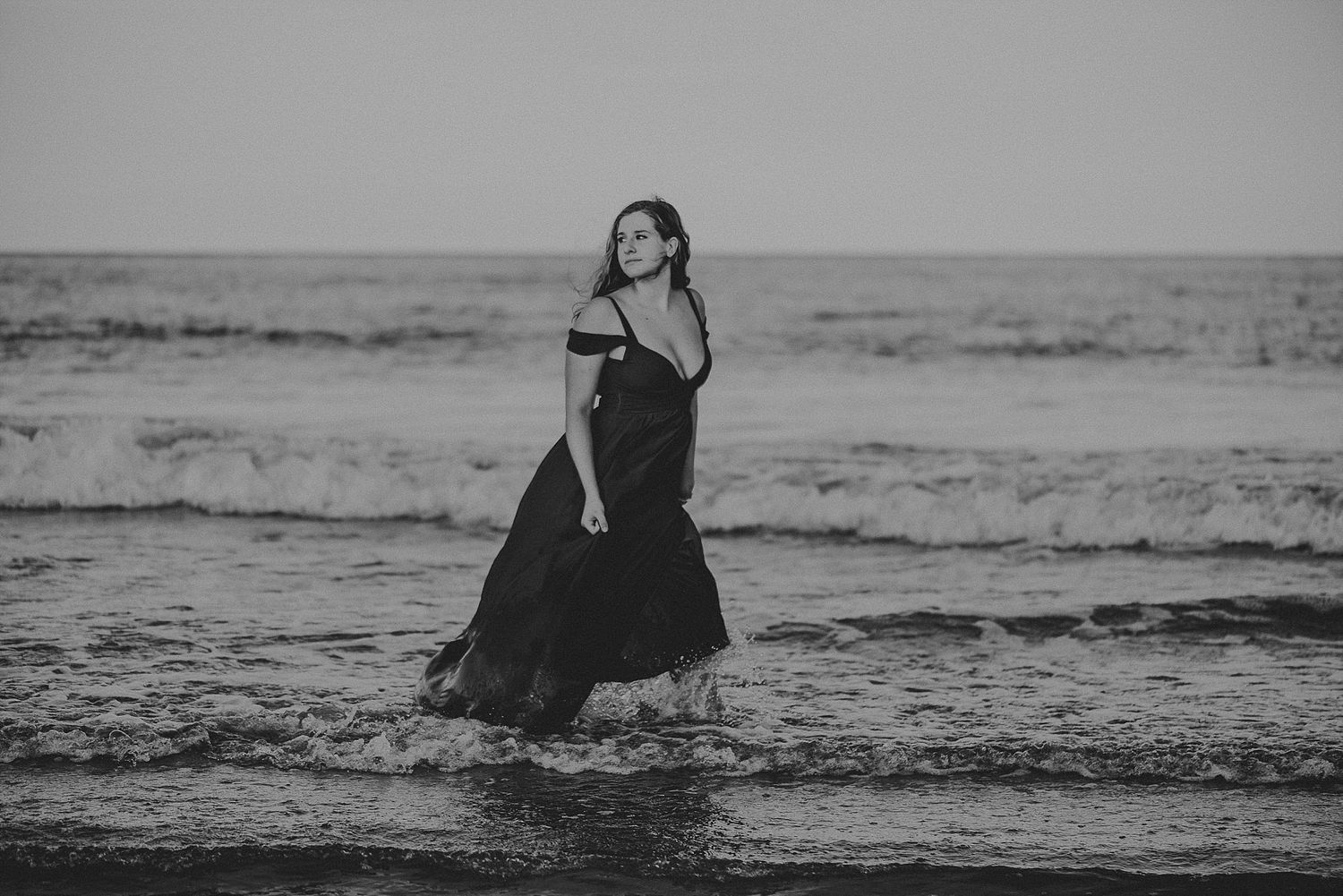 A black dress swirls in the ocean waves during a moody beach photoshoot.