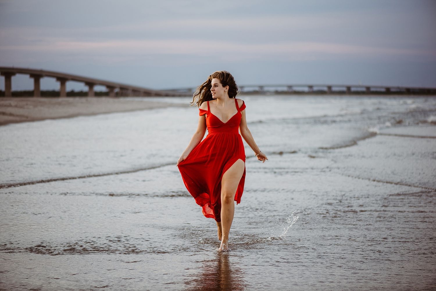 A flowing red dress catches the wind during a beach photoshoot with a bridge in the background.