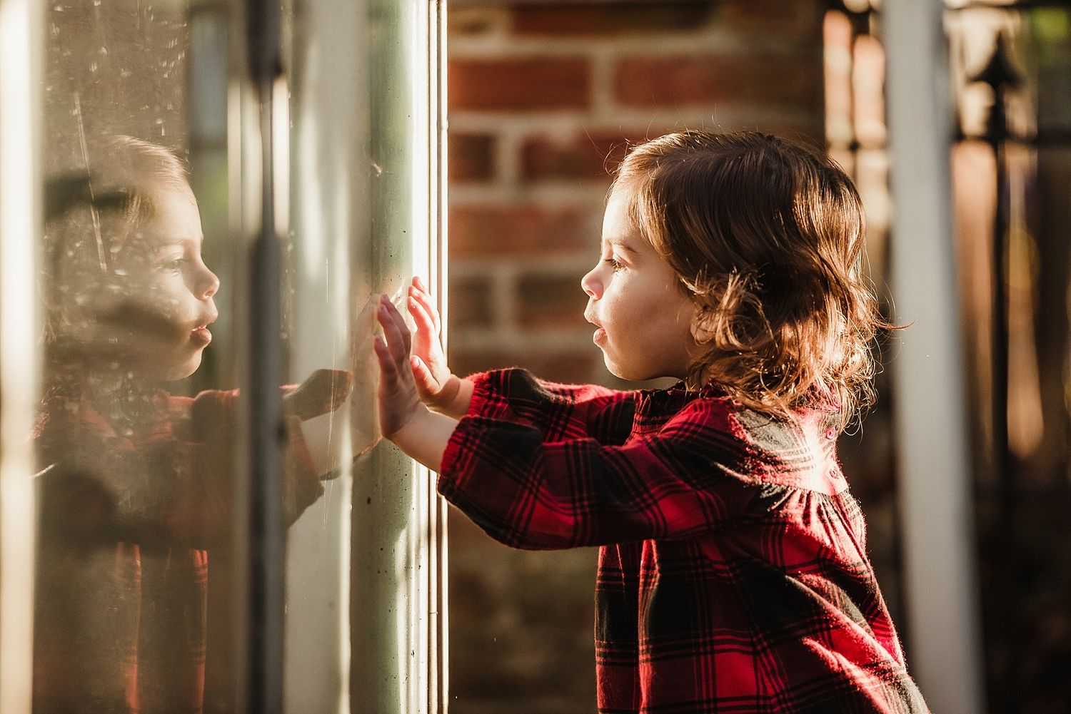 Child in red plaid shirt looking through window reflection in warm sunlit setting.