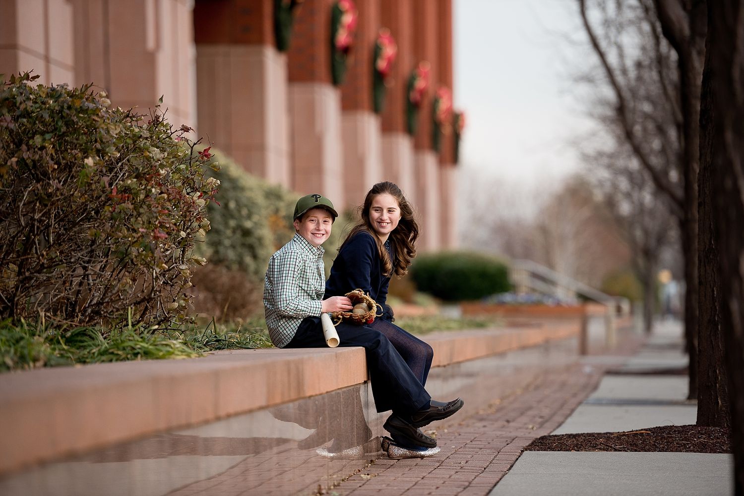 Two people sit together on a brick ledge outside a building with decorative shrubs.