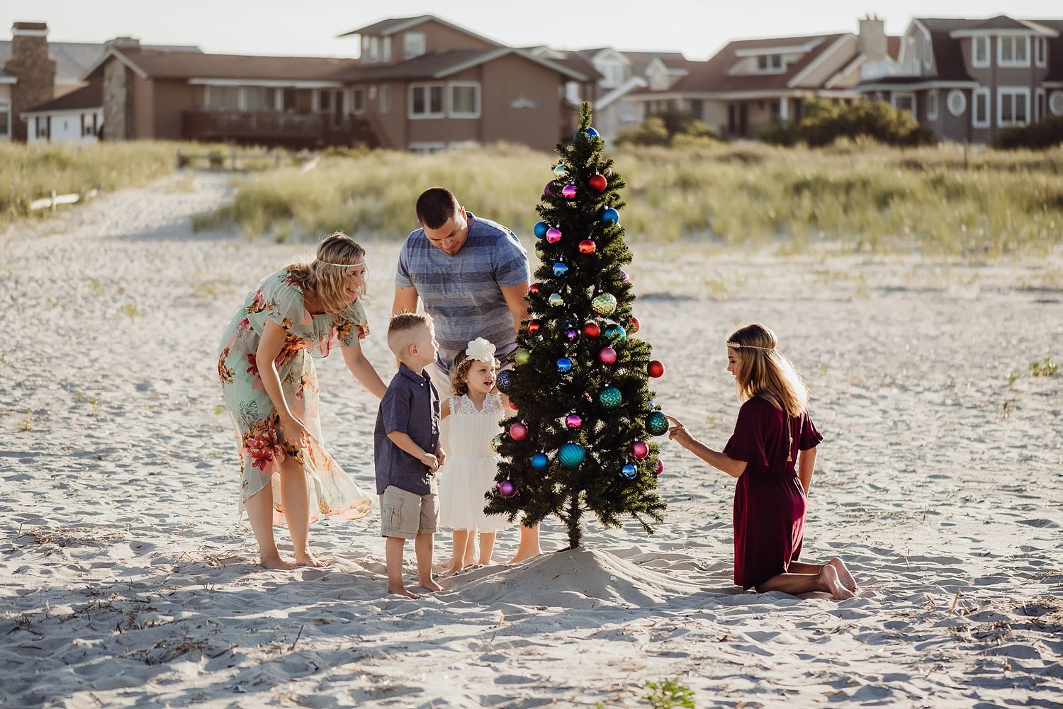 Group decorating Christmas tree together on sandy beach with houses in background.
