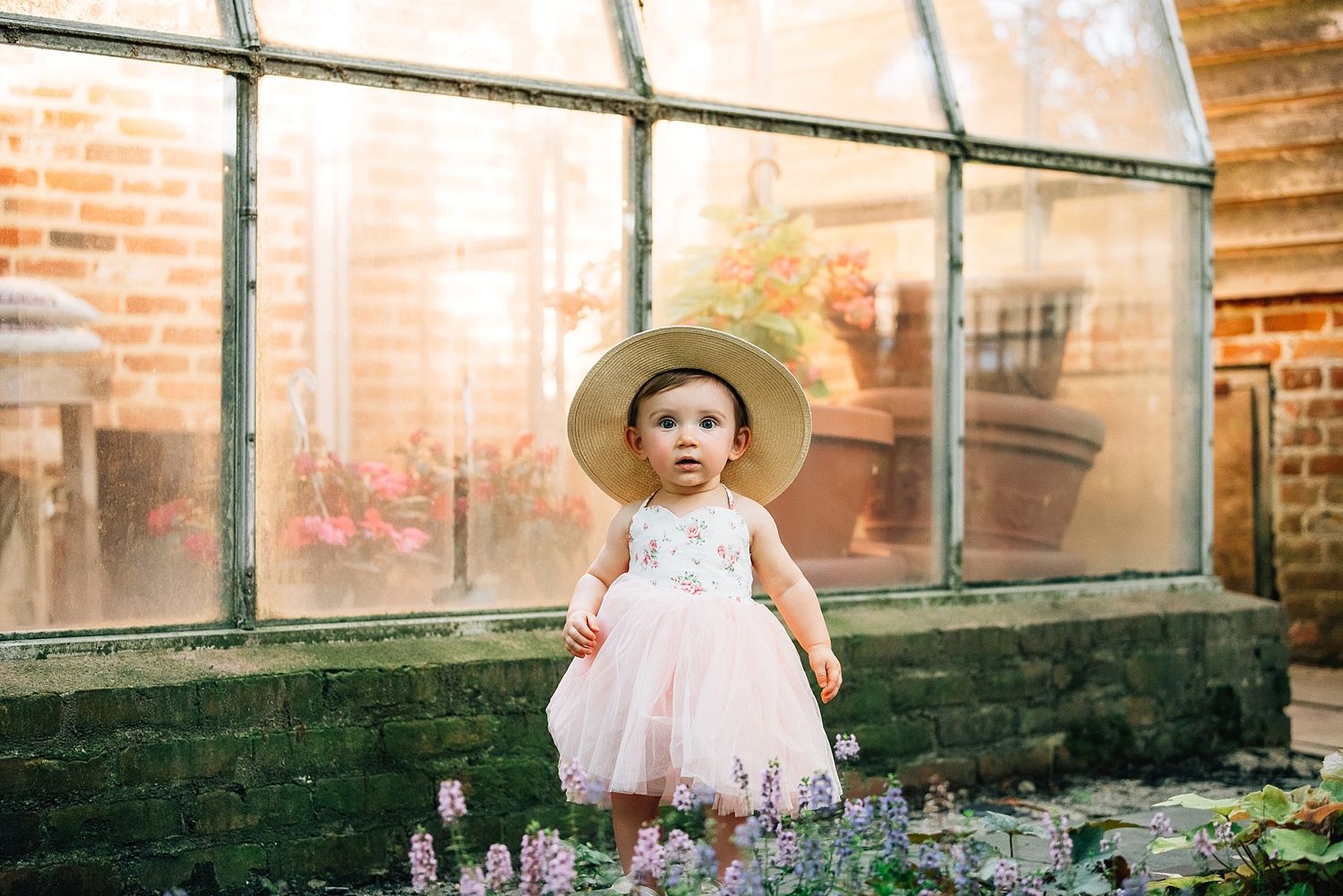 Small child in pink dress and sun hat standing by greenhouse windows.