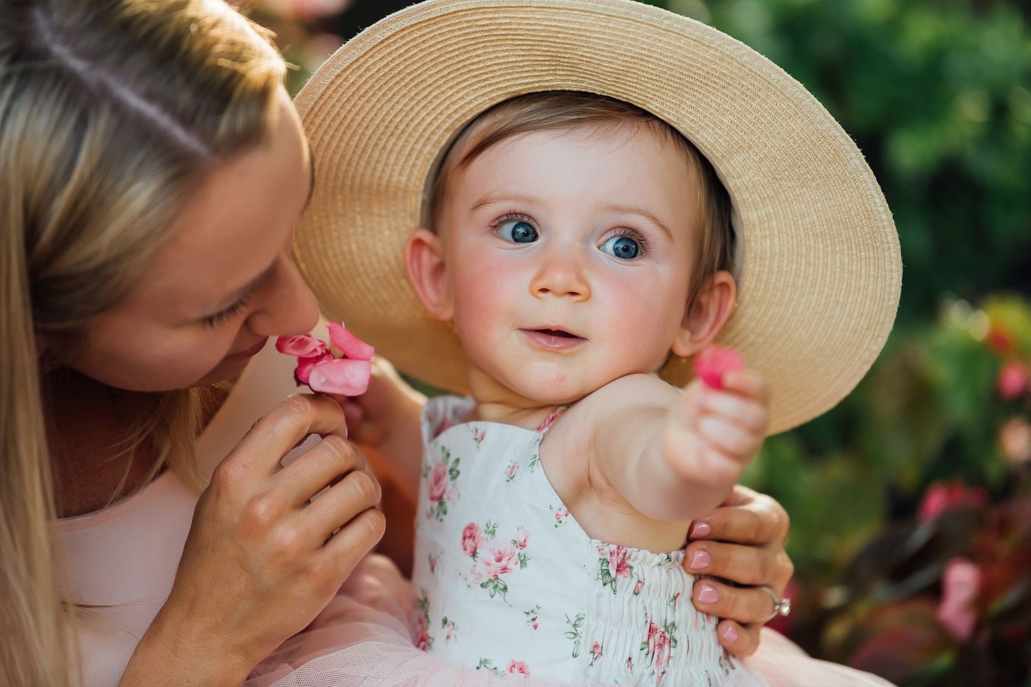 Young child wearing straw hat and floral dress in garden setting.