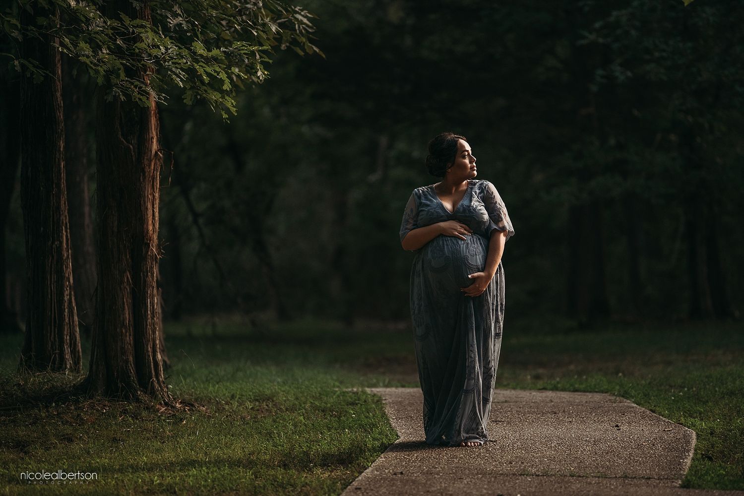 Maternity portrait at sunset on a wooded path shows an expectant mother cradling her belly.