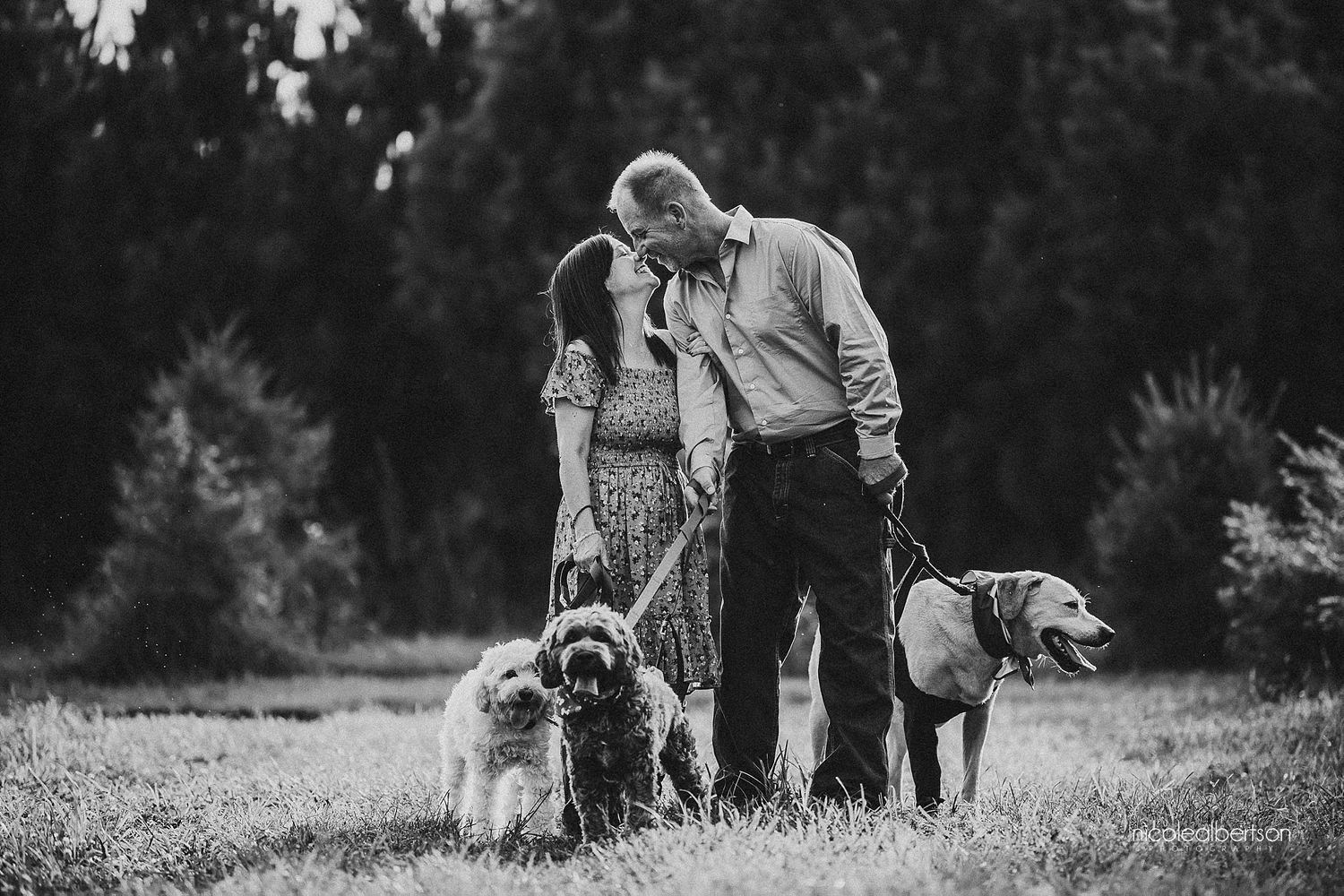 Black and white photo of a couple with their three dogs in a peaceful outdoor setting.