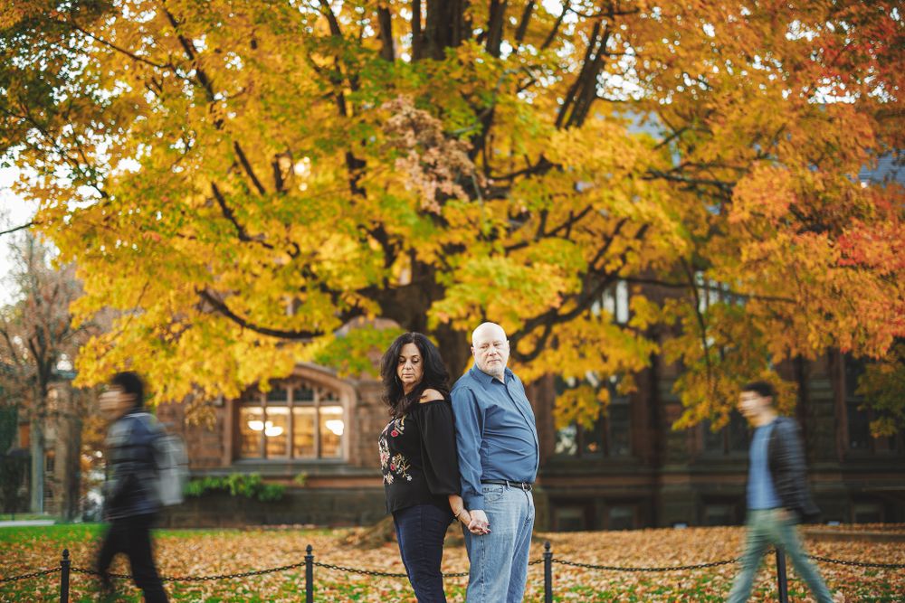 engaged couple standing back to back holding hands serious in front of tree with orange leaves at Princeton University