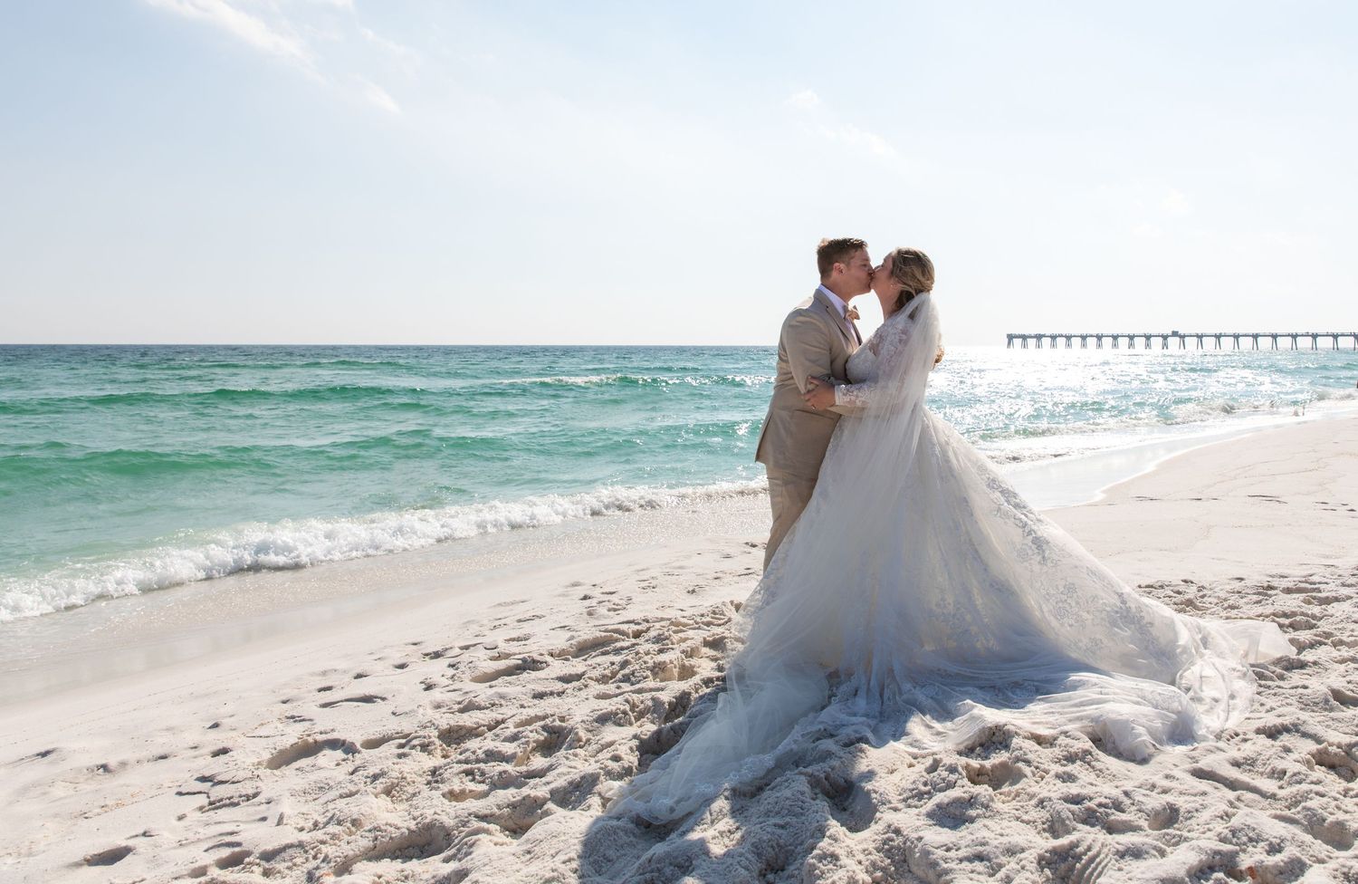 Newlyweds embrace on a white sandy beach with a pier visible in the distance.