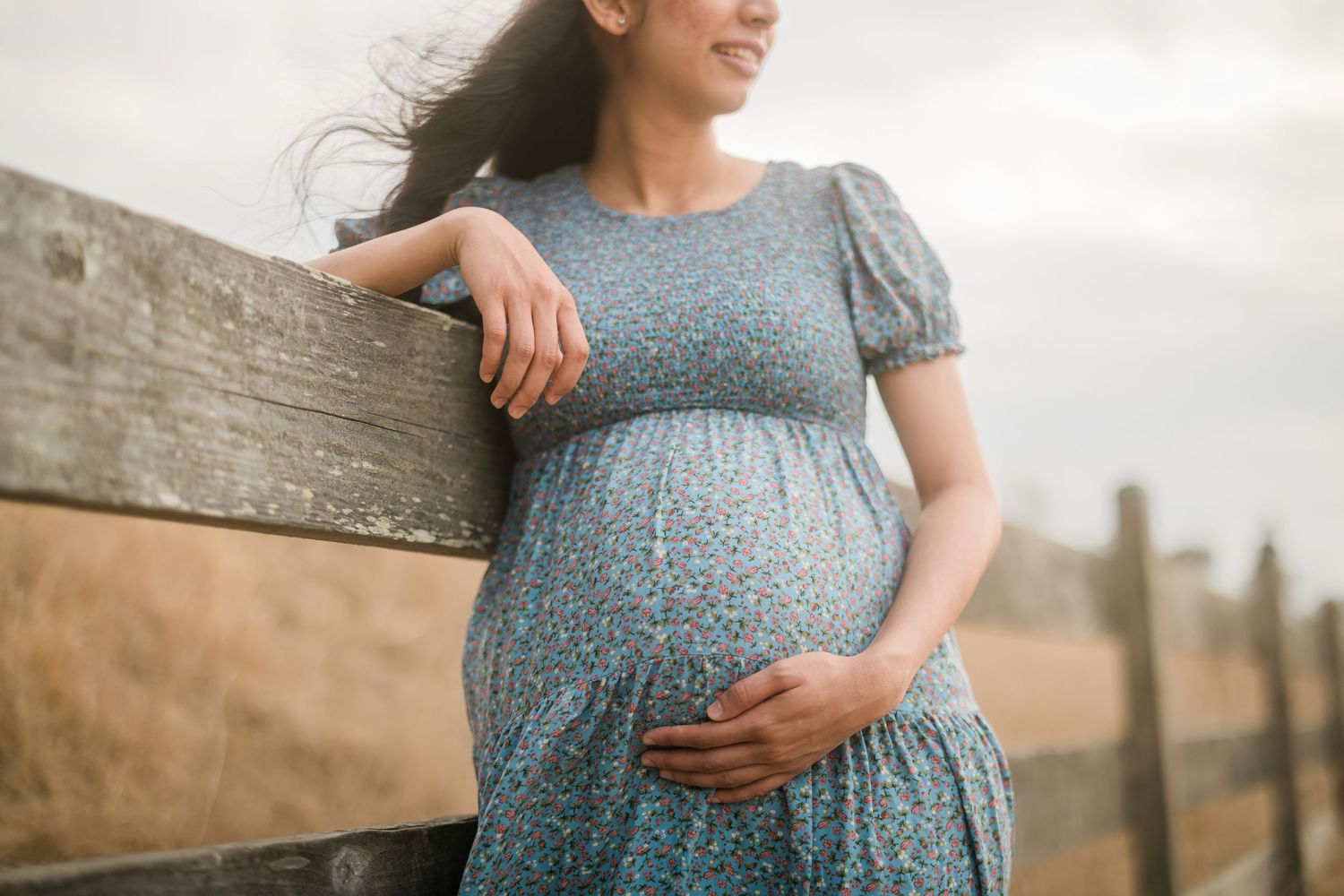 Expectant mother in a blue dress standing by a rustic wooden fence.
