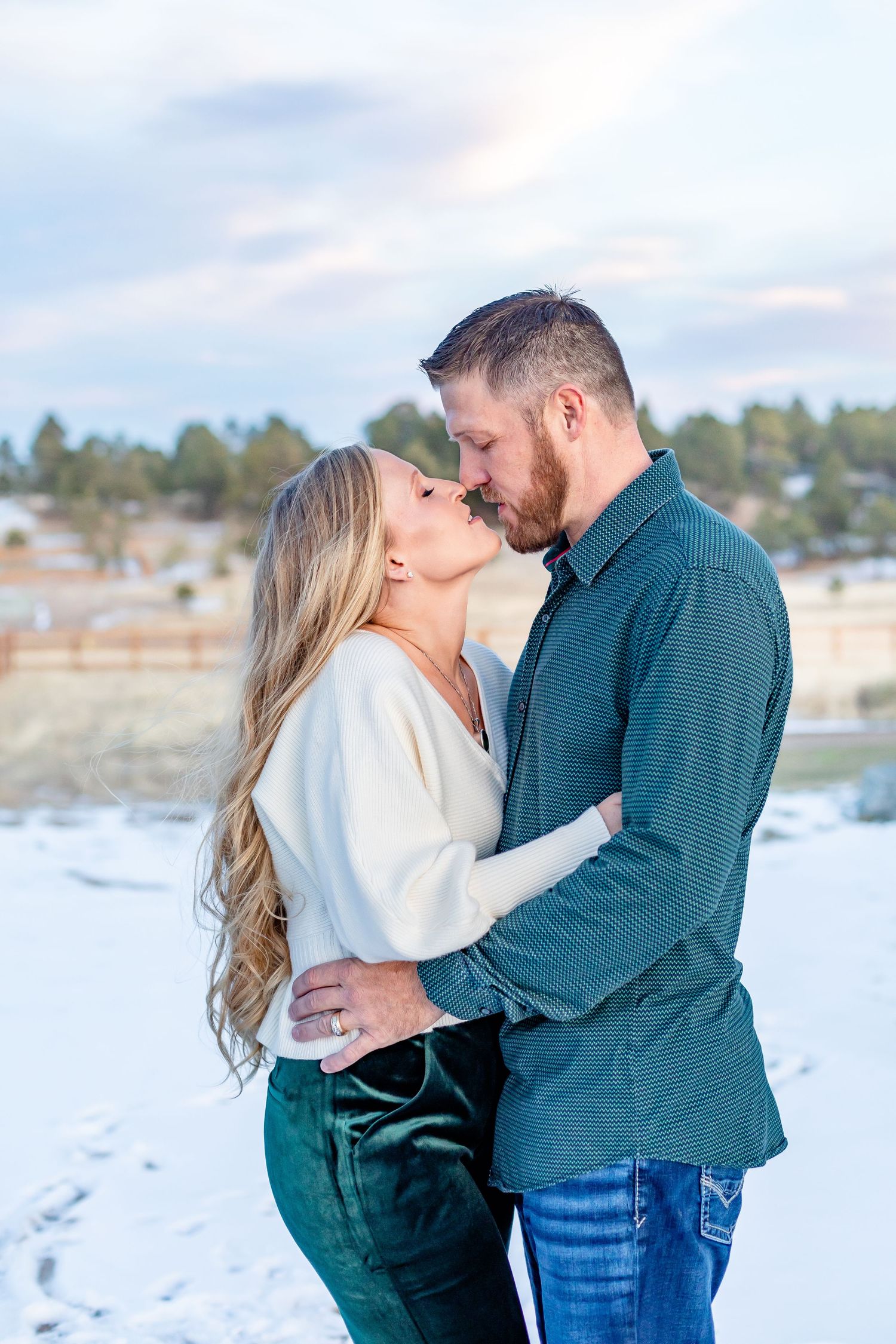 Husband and wife hold each other and move in to kiss while standing in the snow at their property in Black Forest, CO.