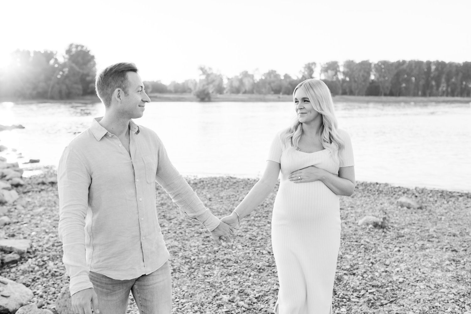 Romantic maternity photo of a couple holding hands by a lake in soft lighting.