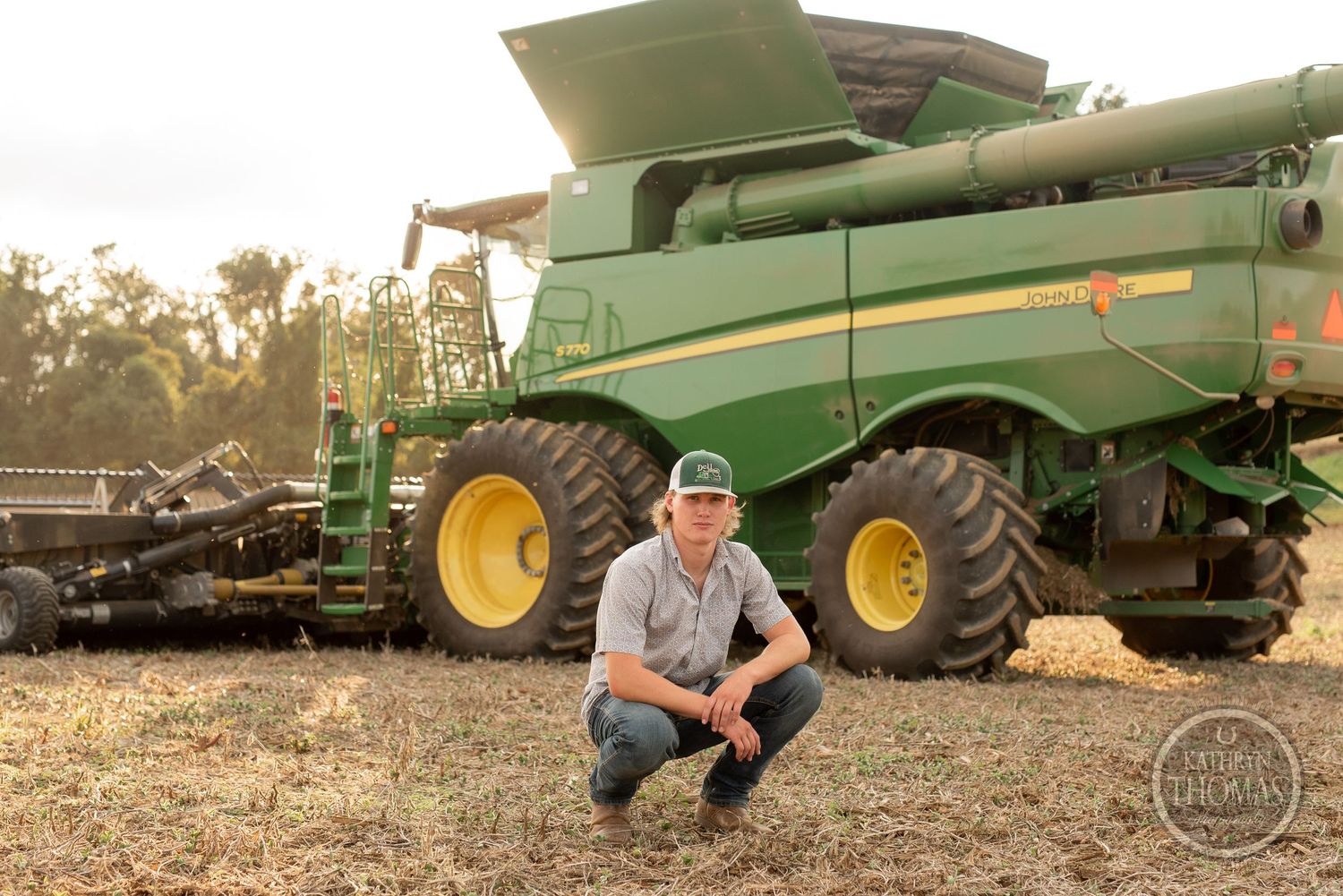 Farmer crouches in field beside large green combine harvester equipment.