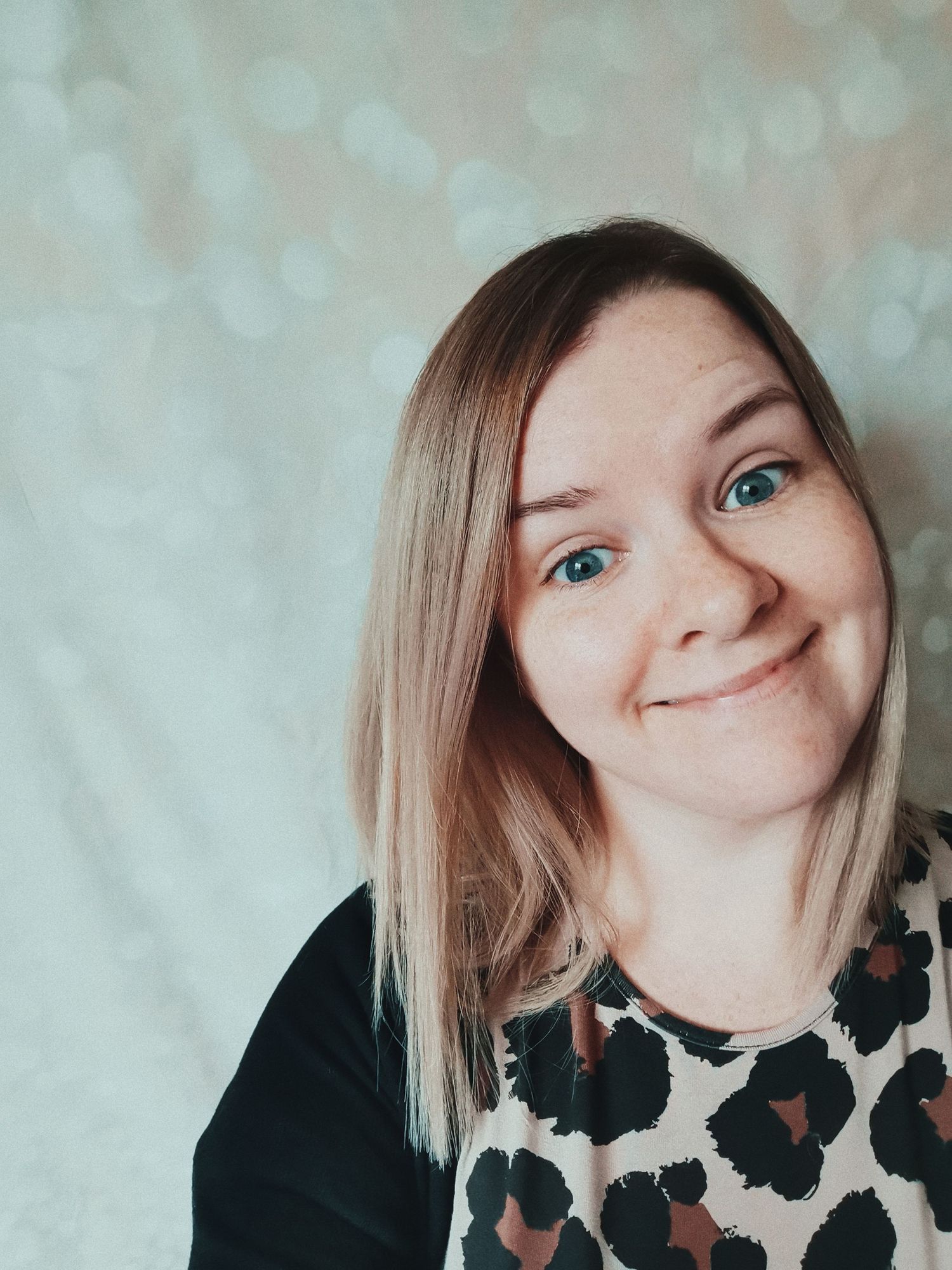 A lighthearted selfie showing bright blue eyes and blonde hair against a soft bokeh background.