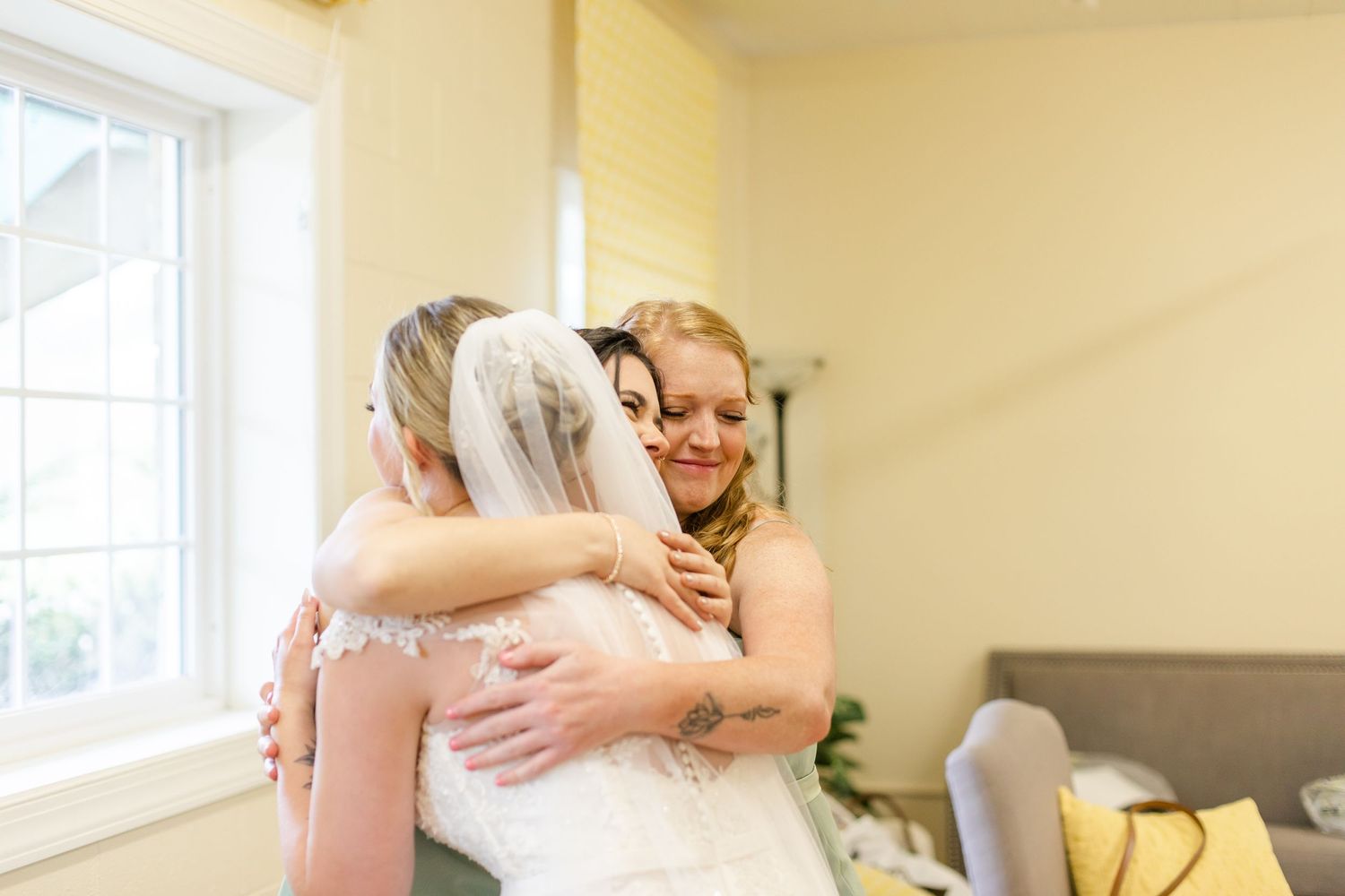 Wedding party members share joyful hugs in a sunlit room with yellow walls.