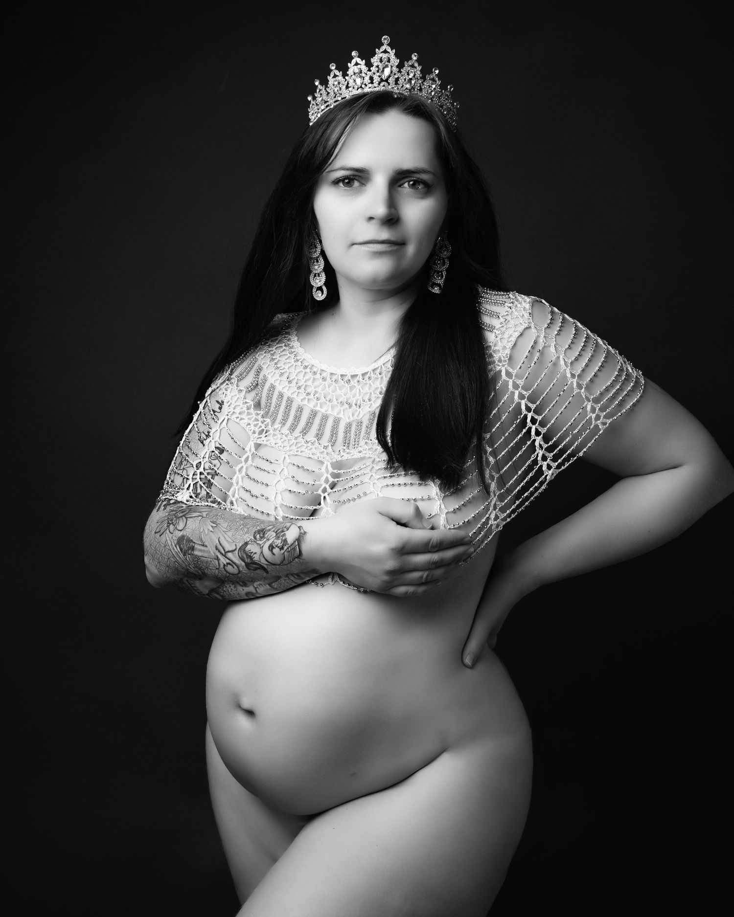 Artistic maternity portrait featuring a woman in a lace top and crown against a dark background.