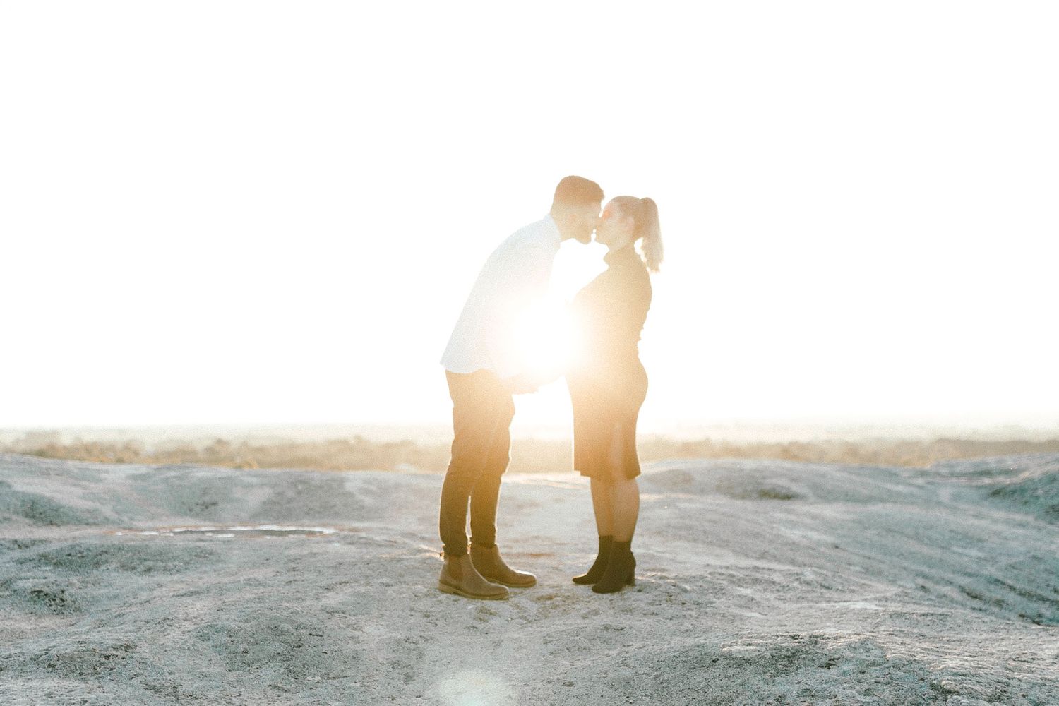 Silhouetted couple stands close together with sun flare behind them on rocky terrain.