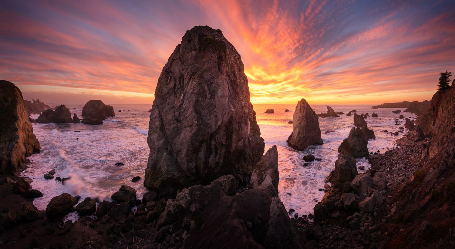 Dramatic purple and orange sunset illuminates rugged sea stacks and rocky coastline with misty waves crashing below.