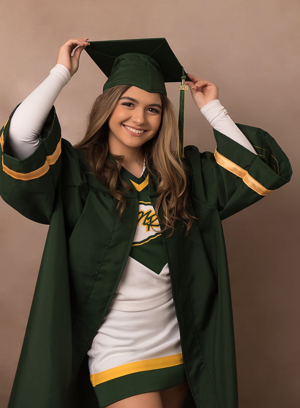 Cheerleader wears graduation cap and gown over uniform in school colors of green and gold.