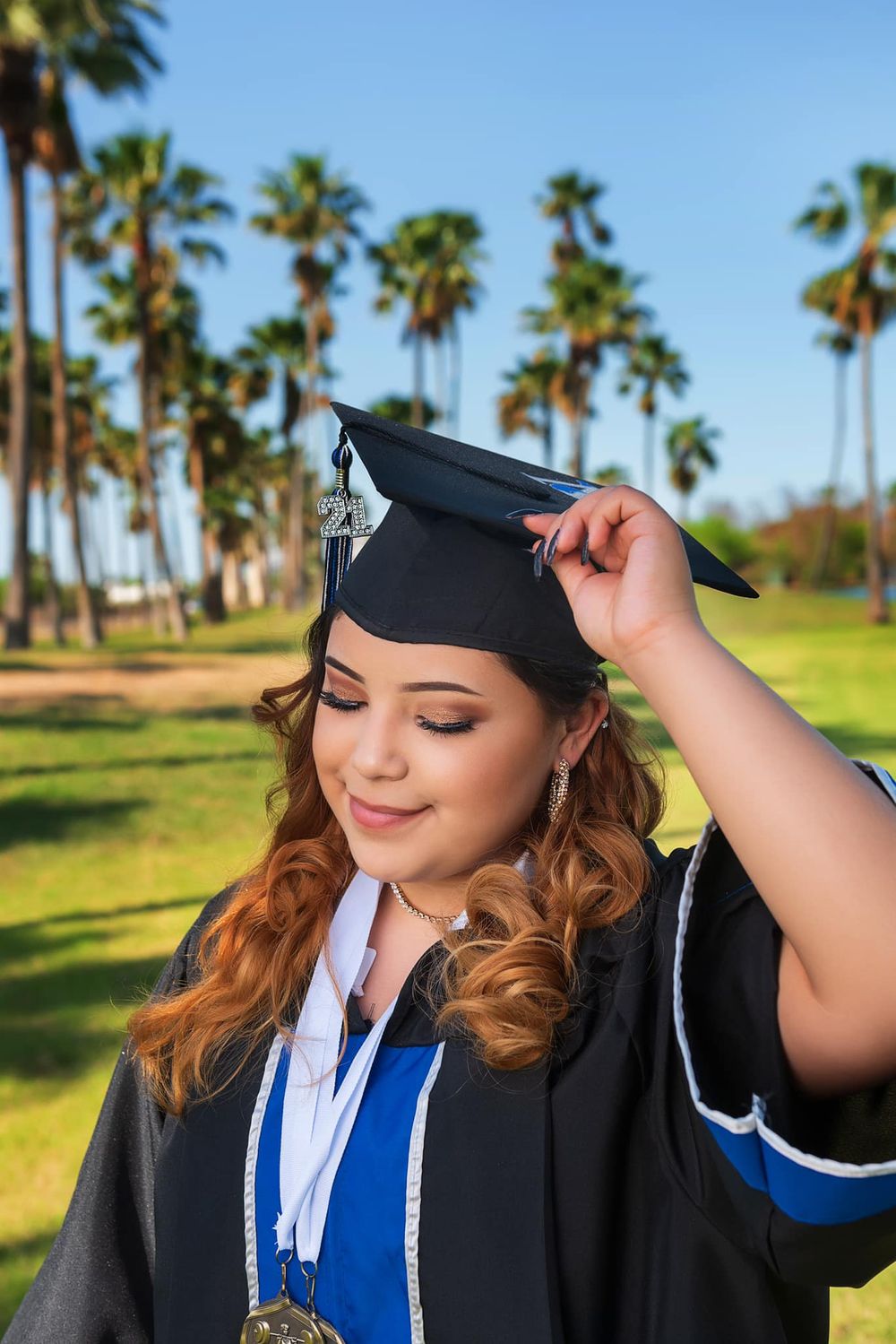 Graduate adjusts mortarboard cap while smiling in sunlit palm tree setting.