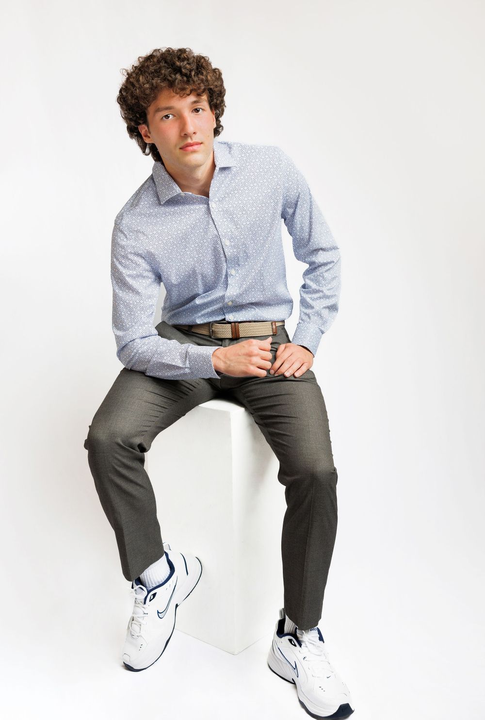 Young man in casual business attire sits on white cube wearing sneakers against white background.