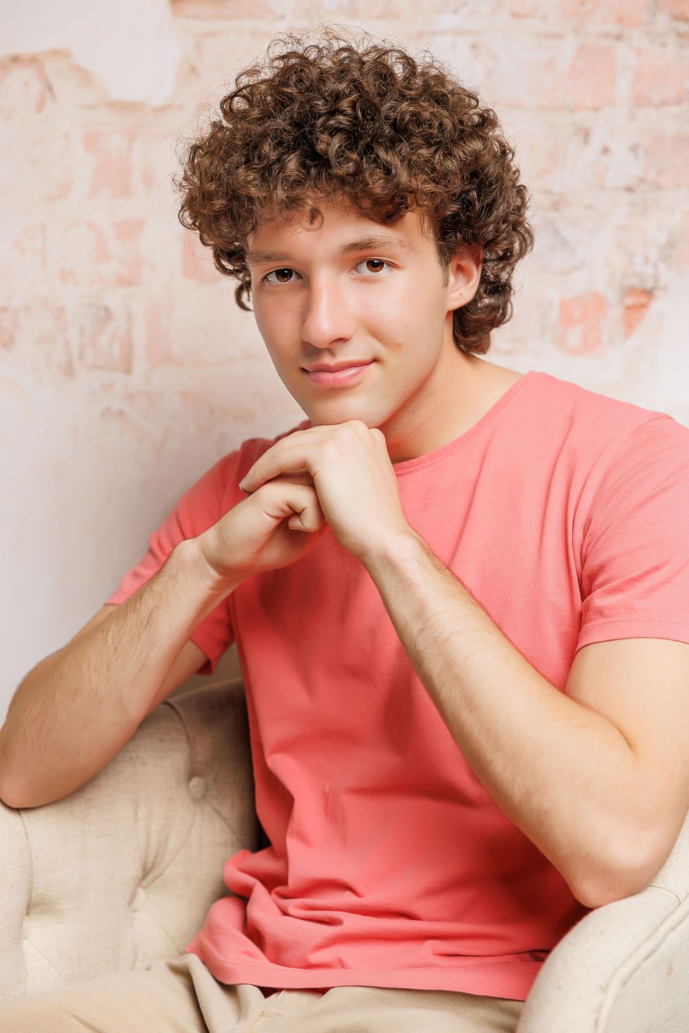 A casual portrait of someone in a coral colored t-shirt with curly hair against a light background.