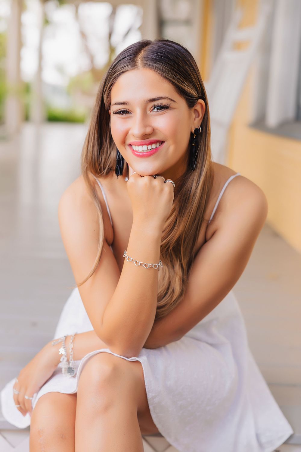 Portrait of someone wearing white dress sitting casually with bright smile and long brown hair against outdoor background.
