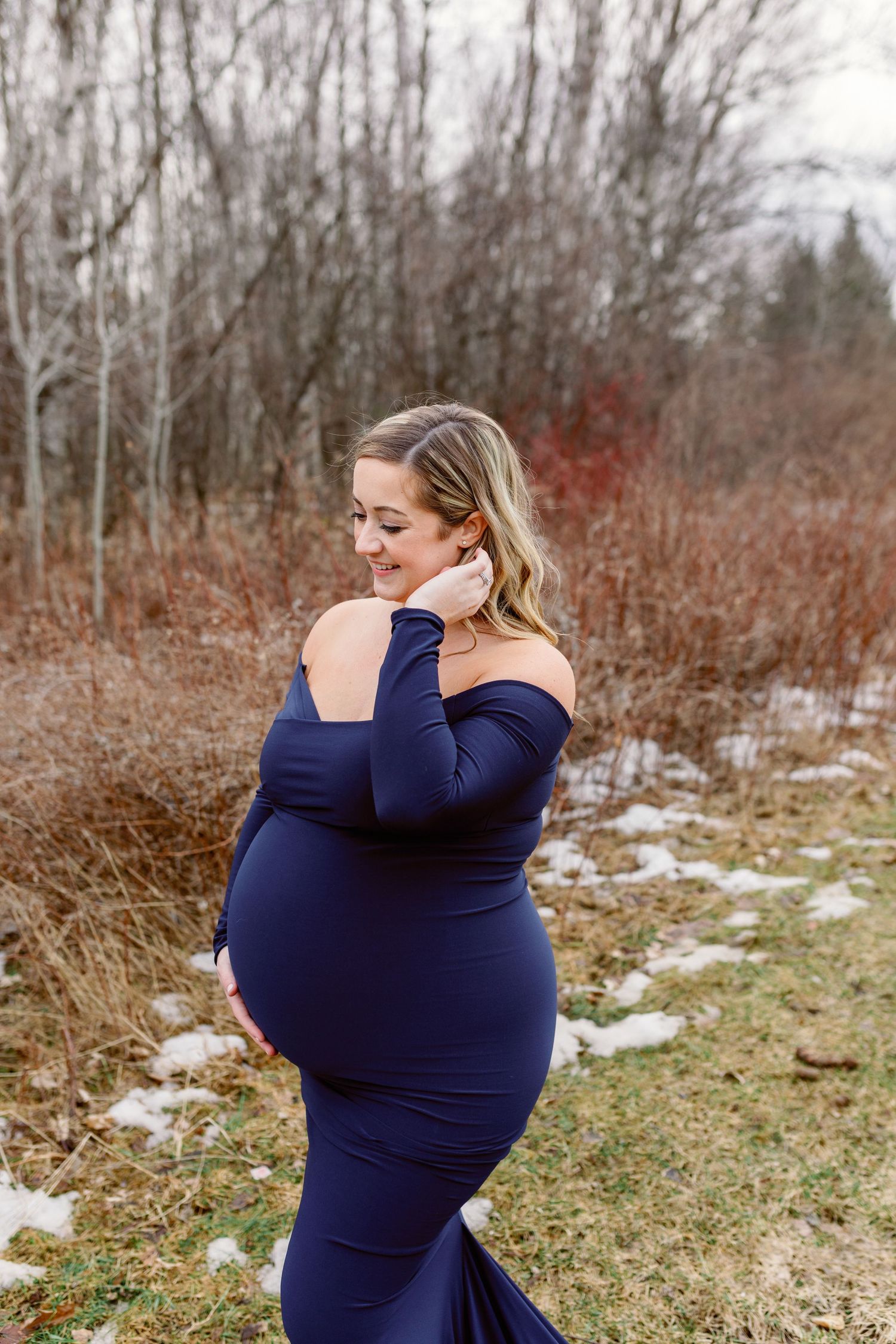A woman in a dark blue dress walks outside, tucking her hair behind her ear, one hand on her belly.  Photo: Katie Crane.