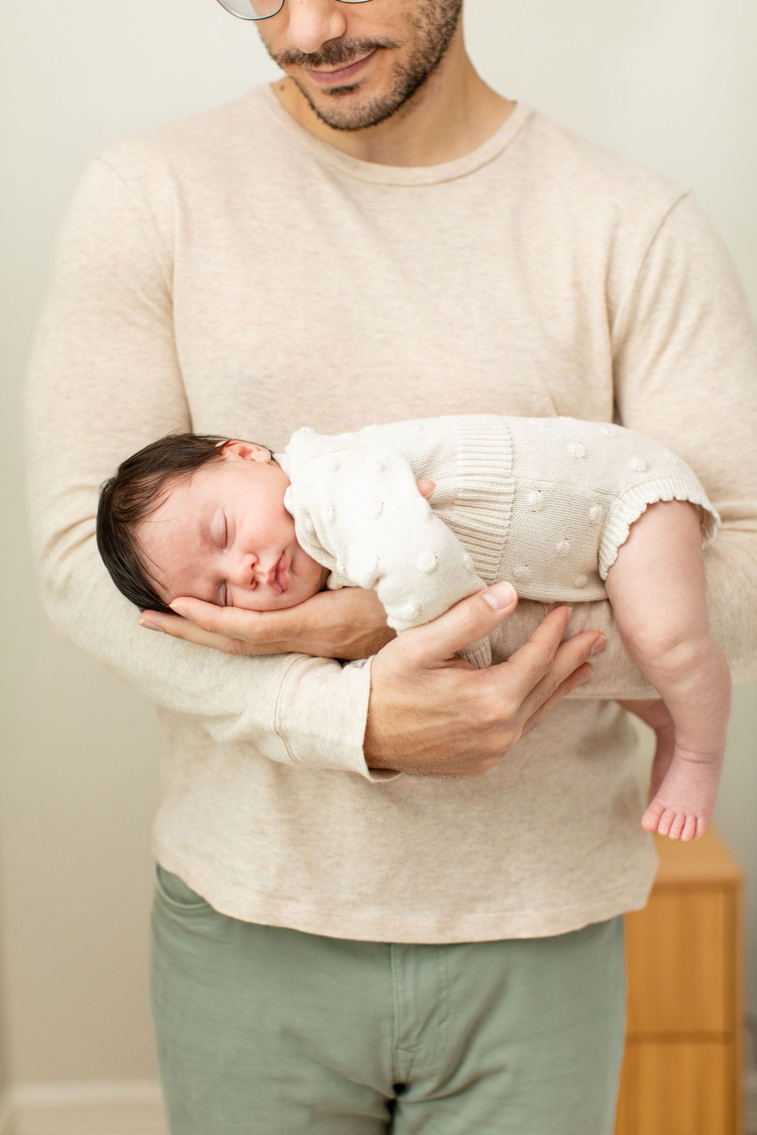 A sleeping baby in white clothing rests in gentle arms against a beige sweater.
