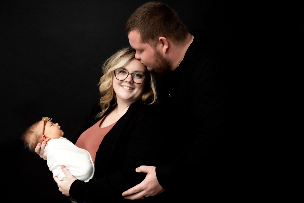 Intimate family portrait against black background showing tender moment between parents and baby.