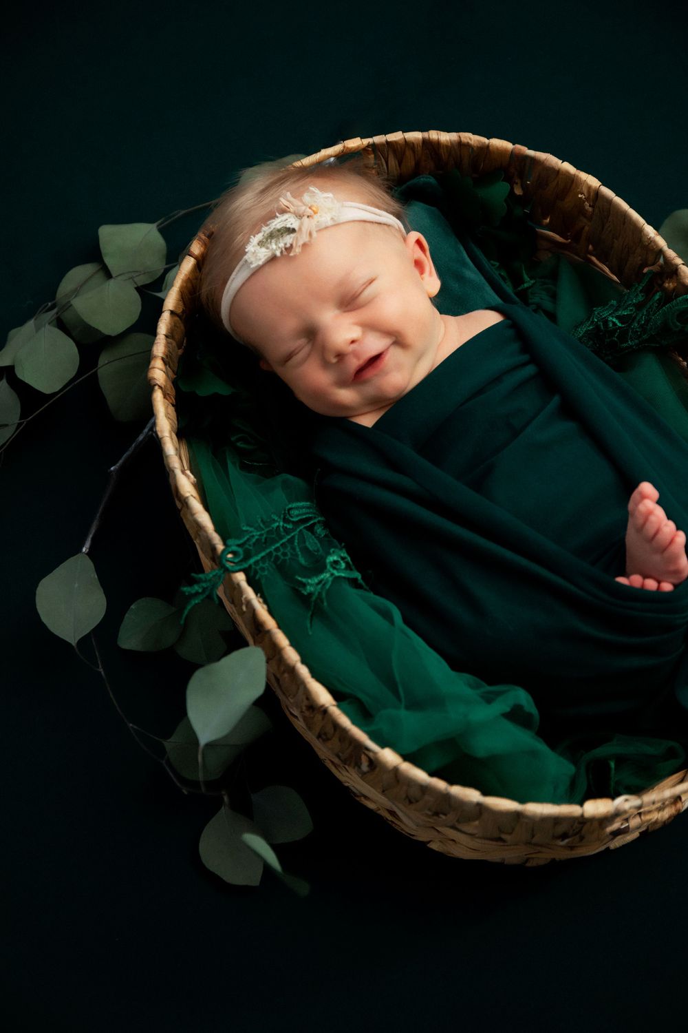 Peaceful newborn wrapped in emerald fabric sleeping in basket with eucalyptus leaves.