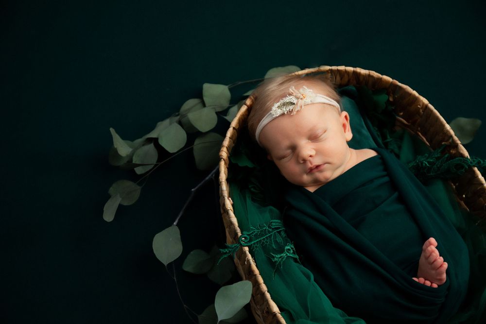 Sleeping newborn in dark wrap nestled in basket with green eucalyptus leaves against dark background.