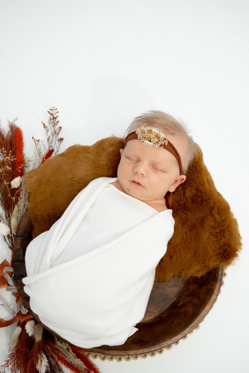 Sleeping newborn in white wrap with delicate headband resting in brown bowl with autumn floral decor.