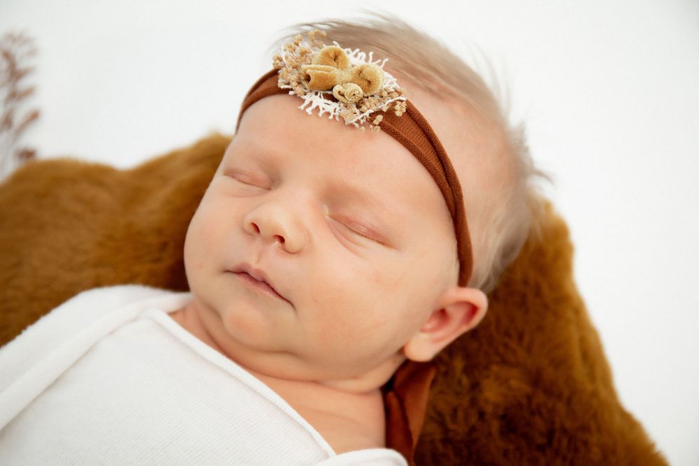 Peaceful newborn wearing ornate headband sleeping in brown bowl with rustic decorative elements.