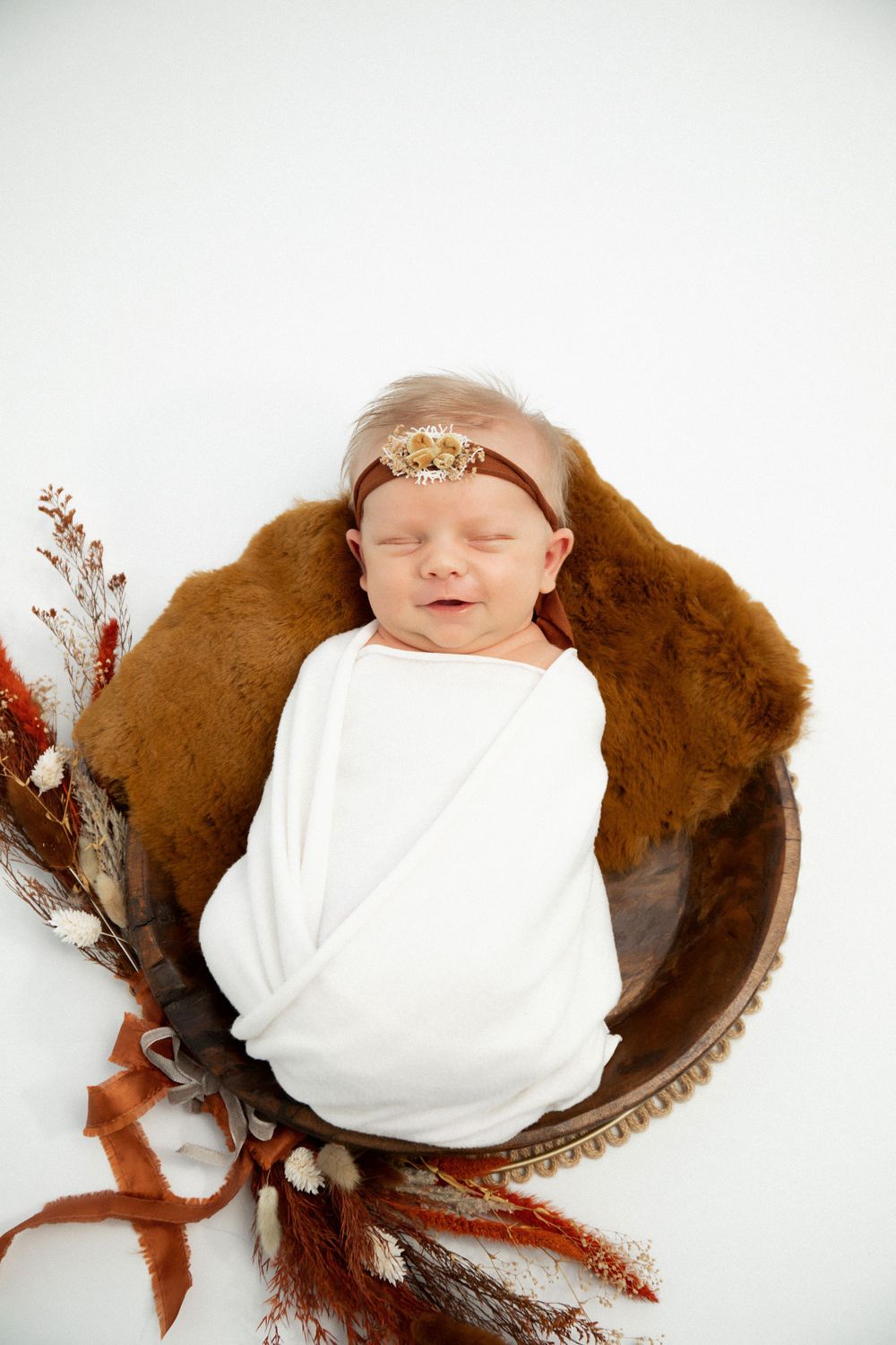 Sleeping baby wrapped in white fabric resting in brown bowl with dried floral accents.