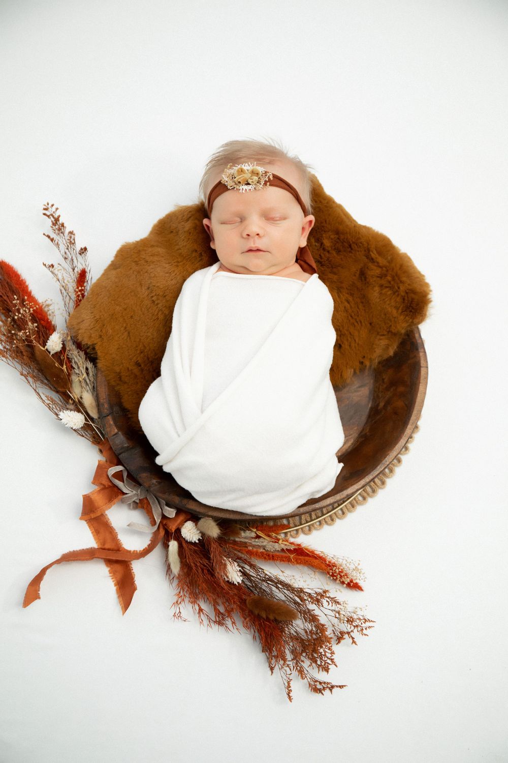 Newborn photography featuring baby in white wrap sleeping in decorative brown bowl with autumn elements.