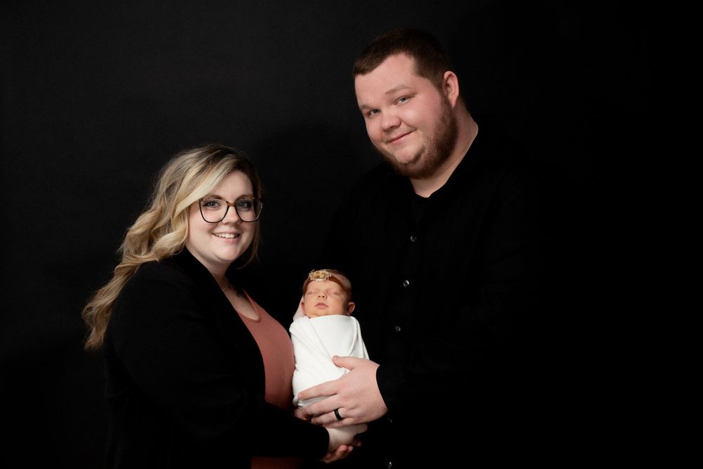Studio portrait of family in black attire against dark background holding newborn.