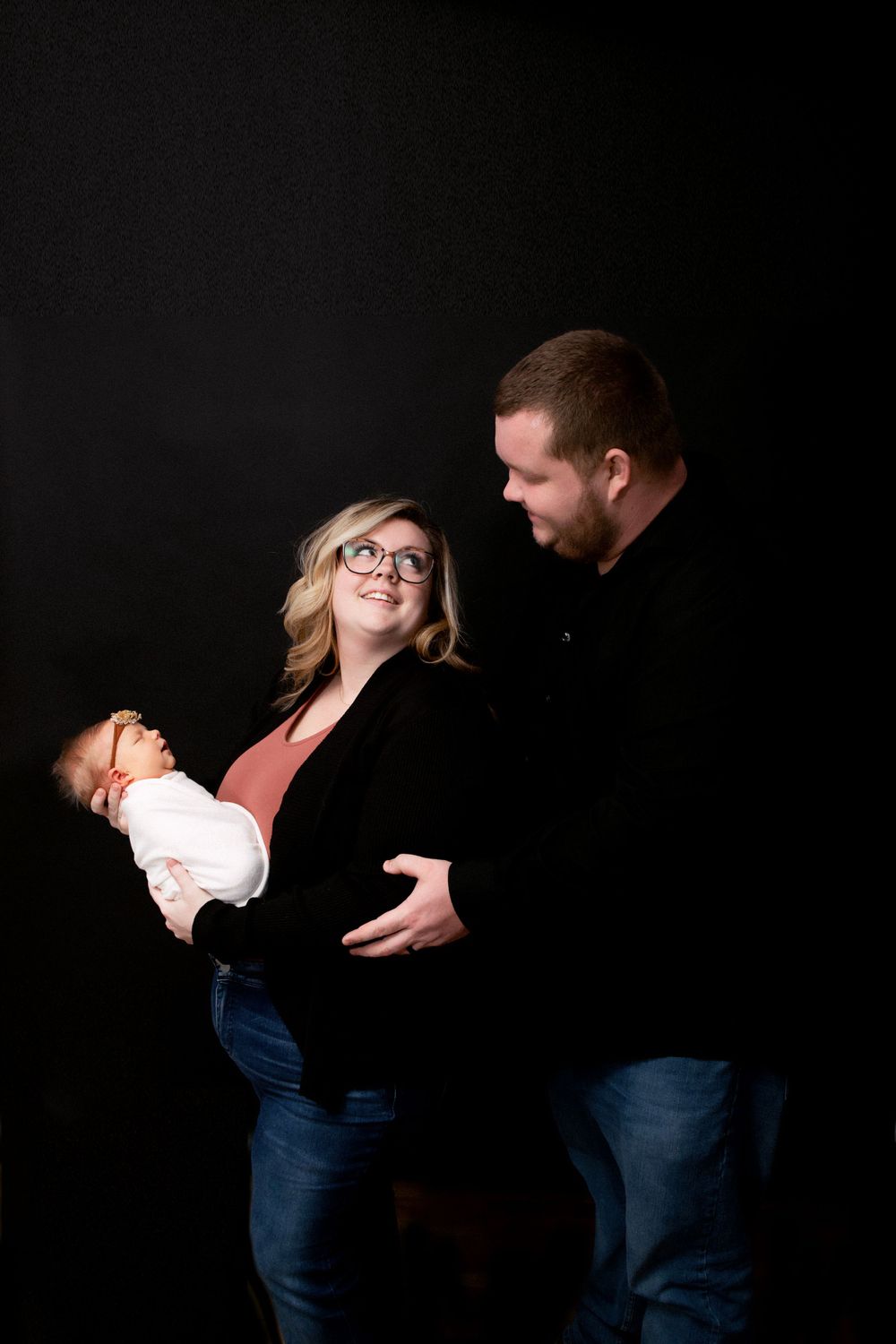 Loving parents in casual wear sharing joyful moment with newborn against dark backdrop.