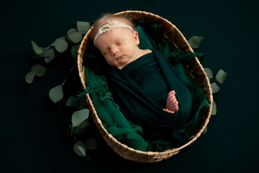 Artistic newborn portrait in green wrap with eucalyptus leaves against dark backdrop.