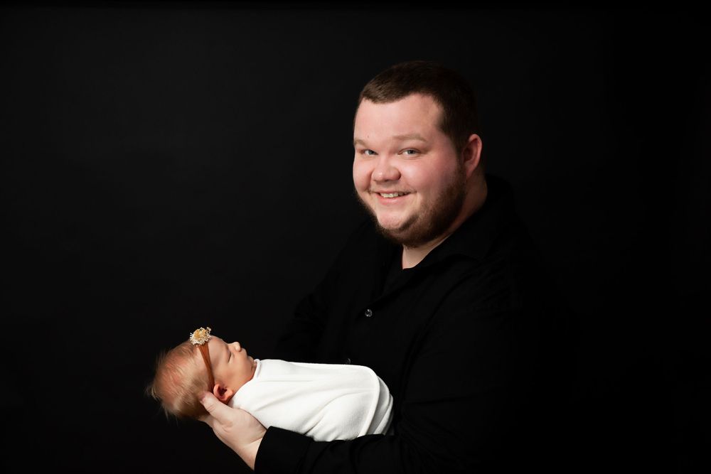 Studio portrait against black background showing parent cradling infant in white outfit.