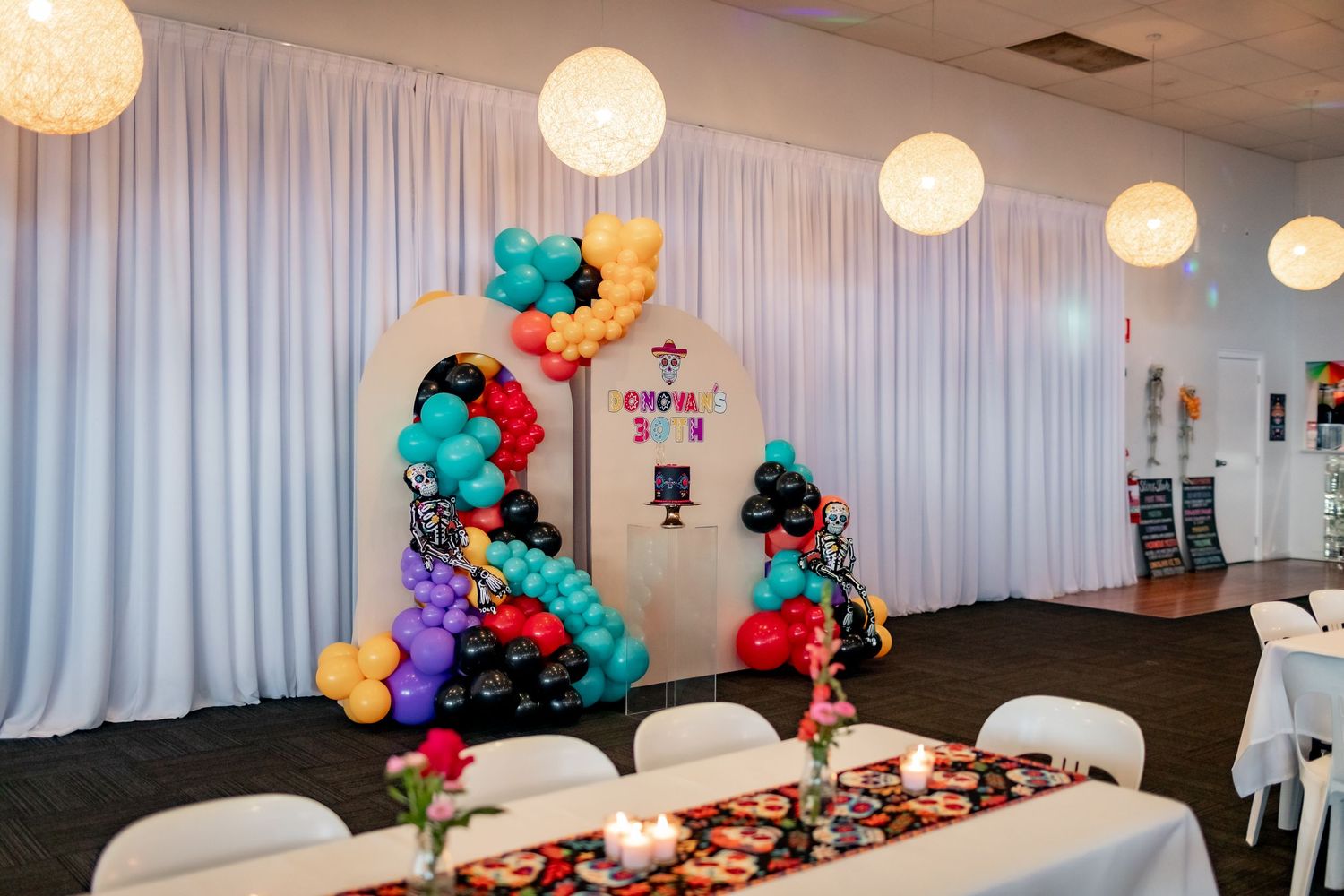 A festive party setup with colorful balloon arch and white draping over dining tables.