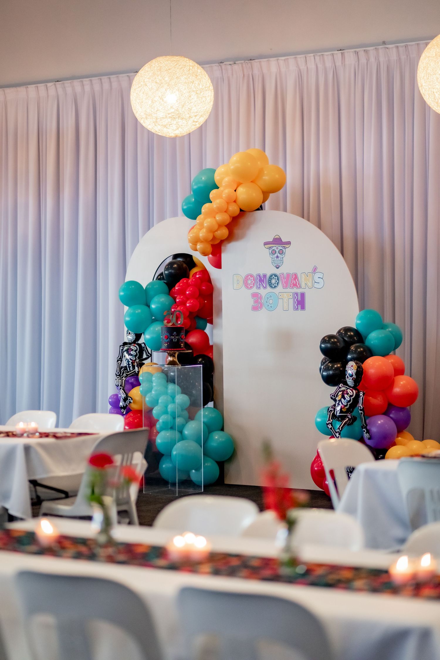 A white decorative arch adorned with vibrant balloon garlands and hanging globe lights.