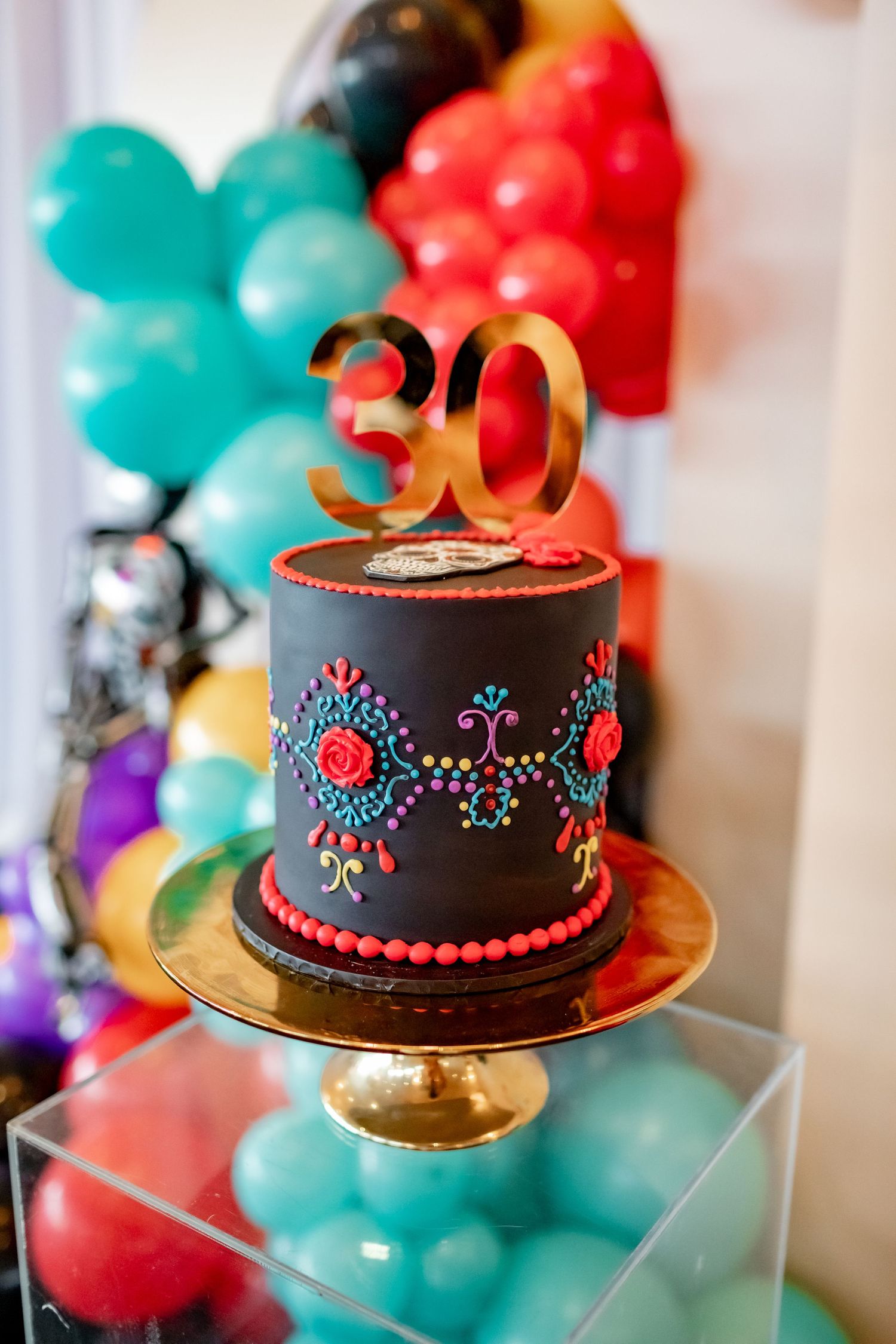 A black fondant birthday cake decorated with colorful floral designs and topped with a gold number 30 on a gold cake stand.