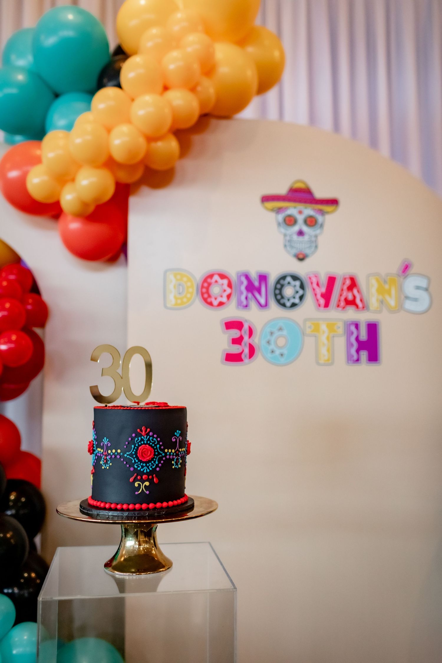 A black birthday cake with colorful details under a Day of the Dead themed party backdrop.