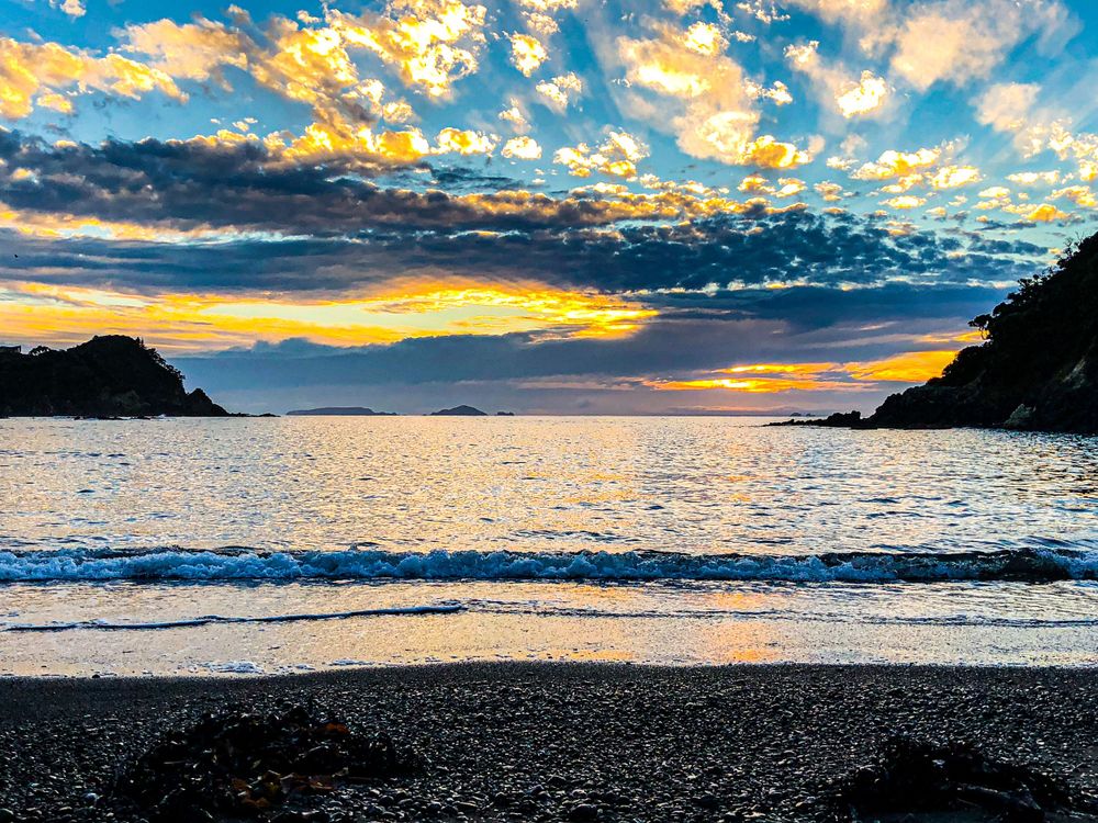 Vibrant sunset sky with golden clouds reflects over calm ocean waters and dark sandy beach.
